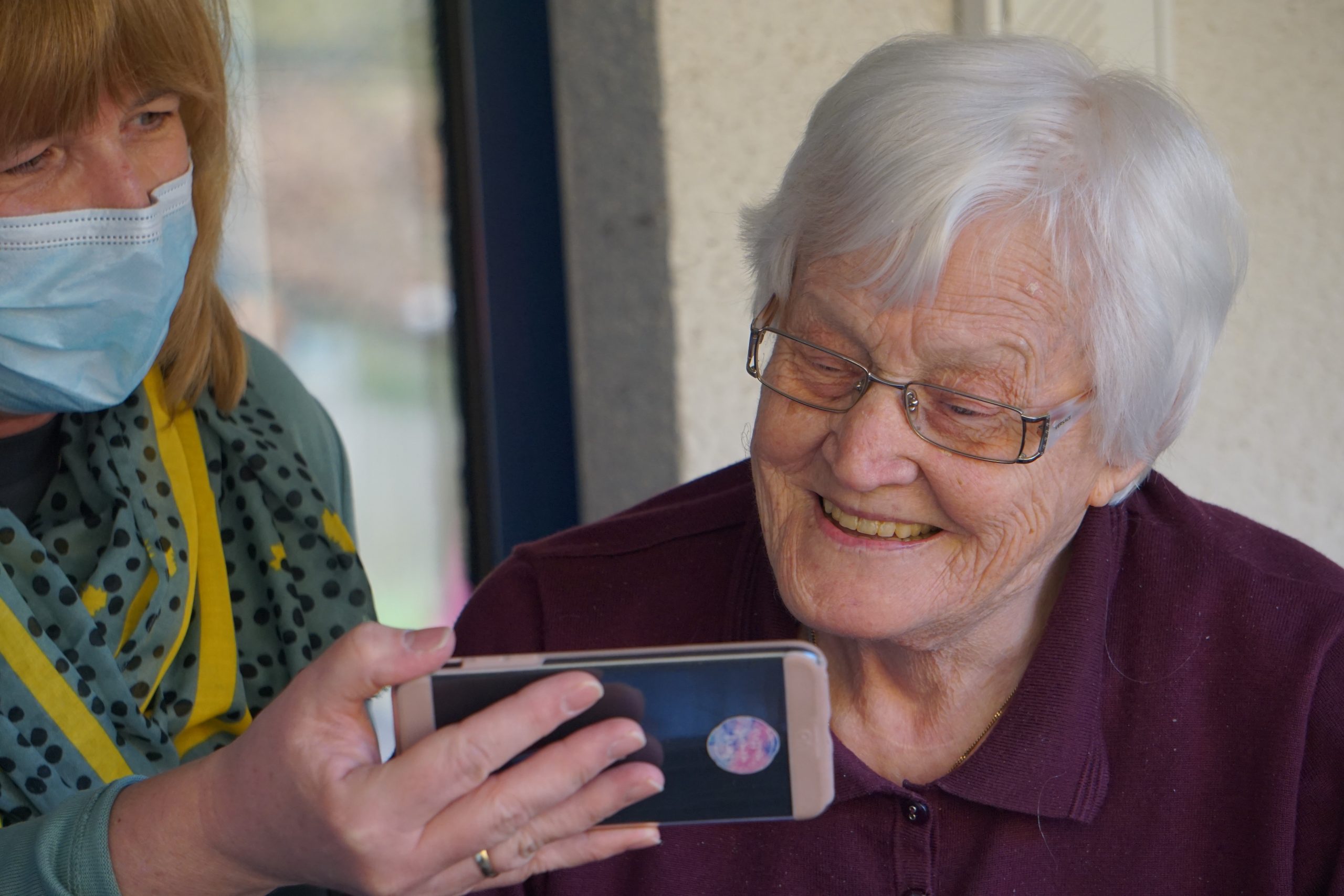 A woman wearing a mask help an elderly woman use a mobile phone